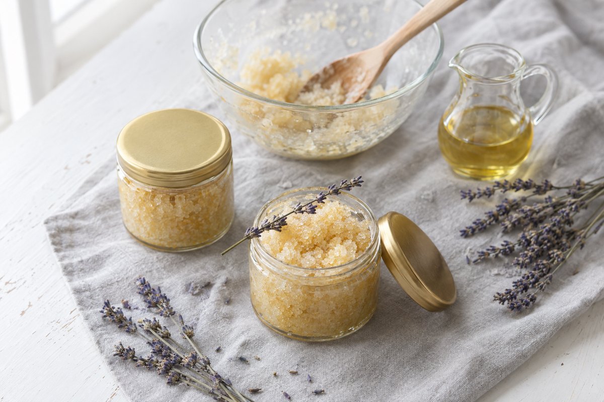 Two small glass jars of pale golden homemade sugar scrub with gold lids beside dried lavender sprigs on grey linen